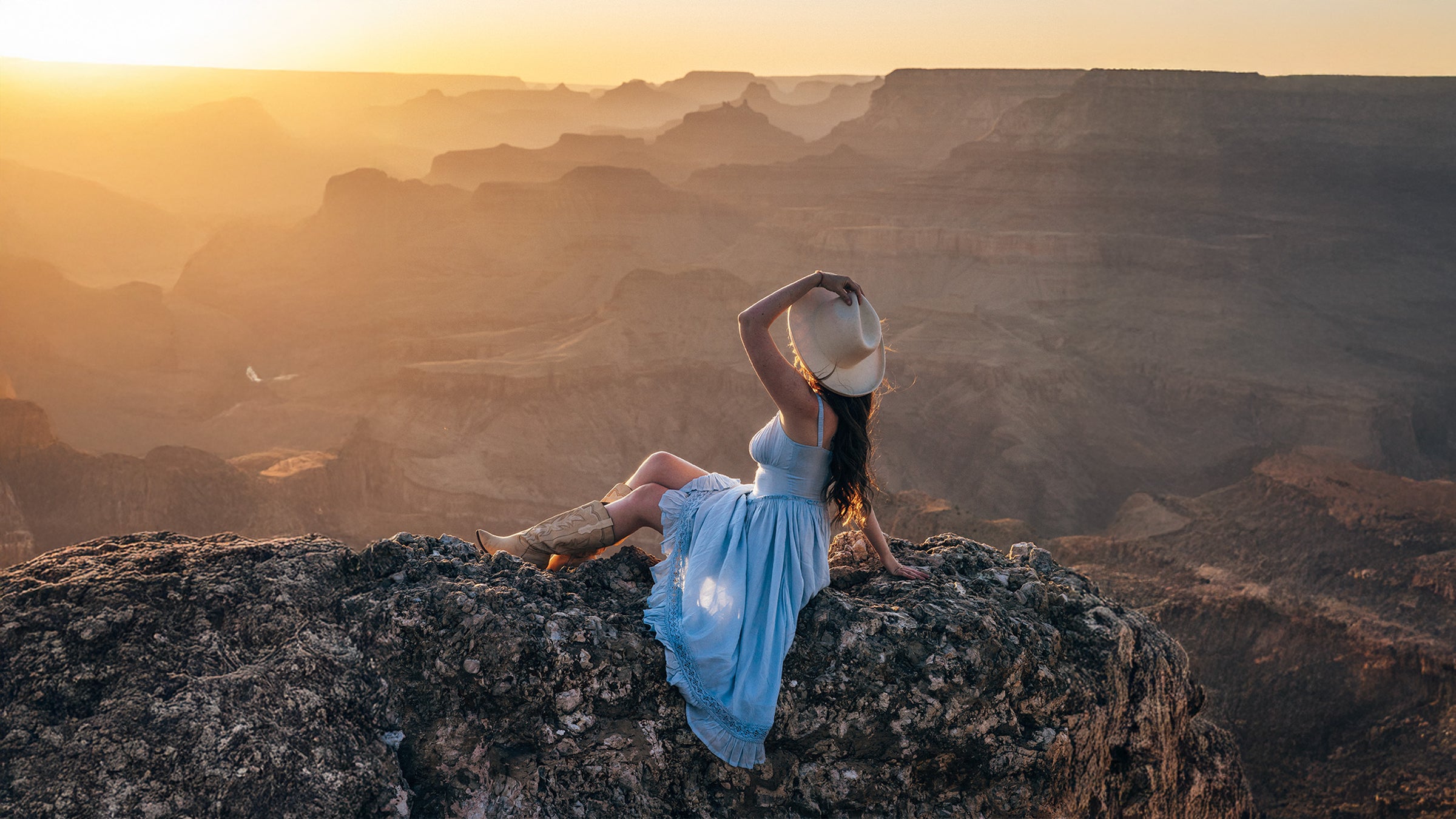 Woman in a blue dress sitting on a rocky outcrop overlooking a vast landscape at sunset.