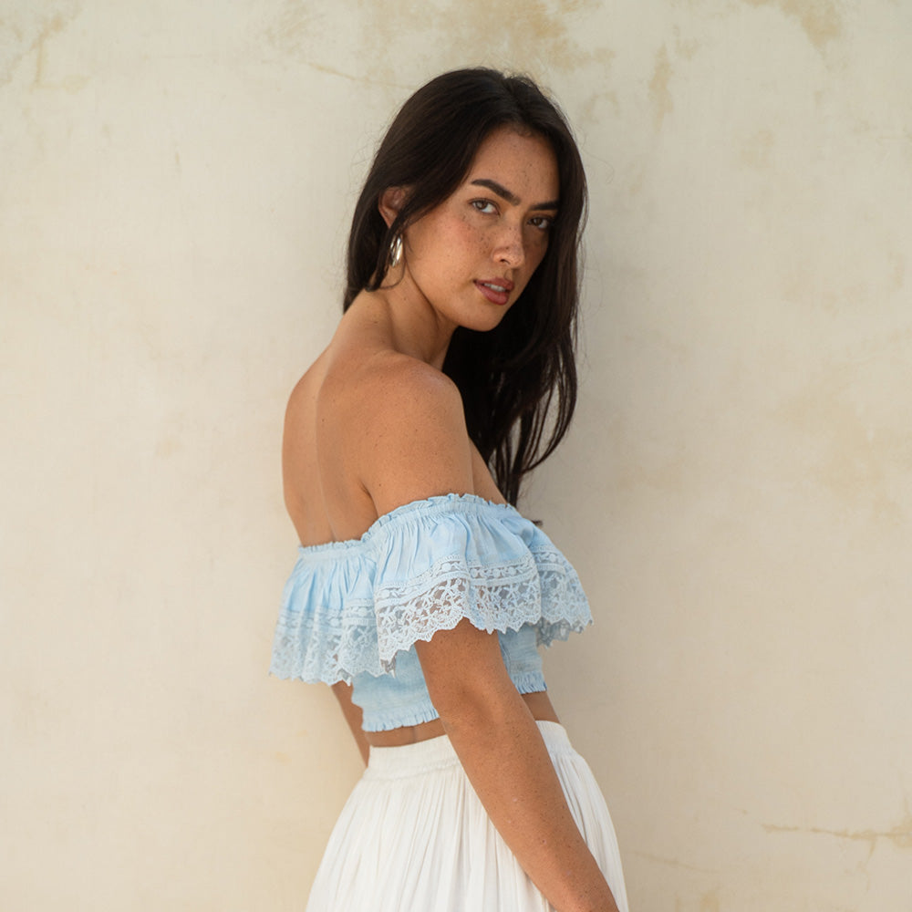 Woman wearing a light blue off-shoulder top with lace details against a beige wall.