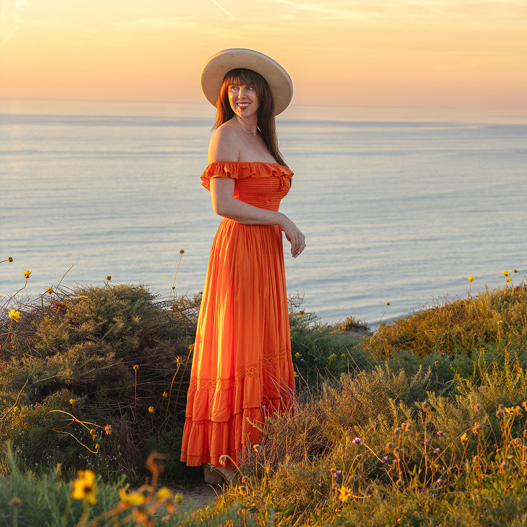 Woman in an orange dress and hat standing by a body of water at sunset.