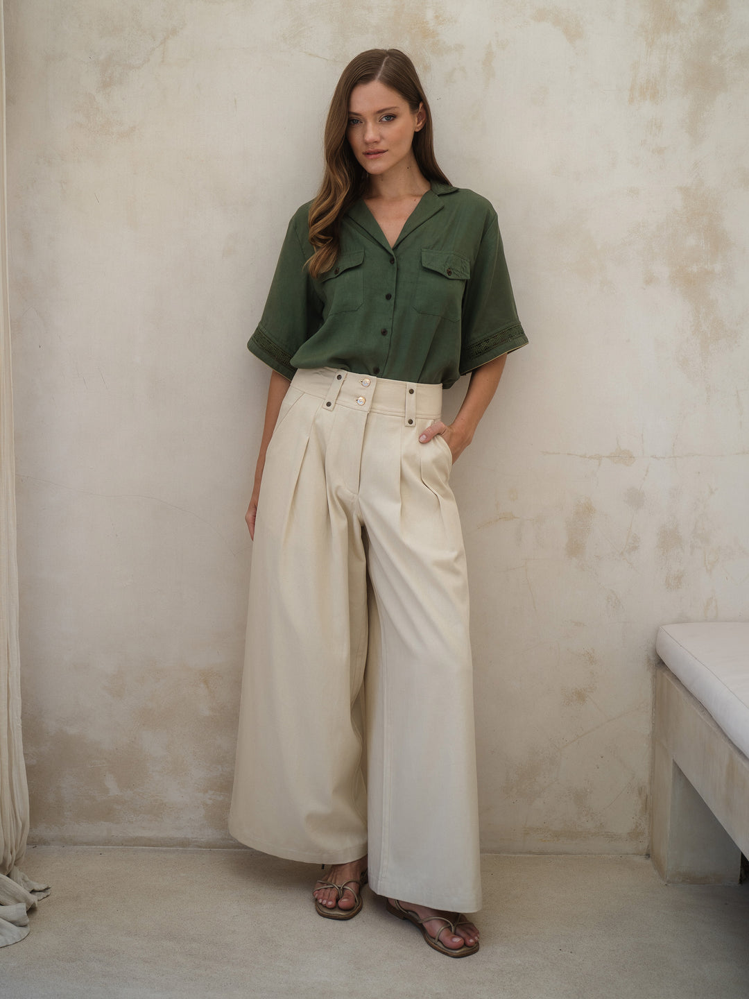 Woman wearing a green shirt and beige pants against a plain wall.