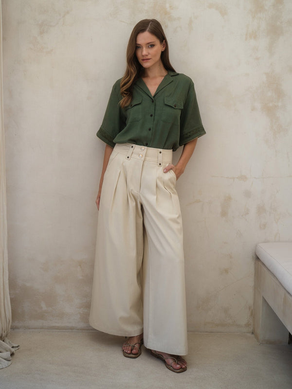 Woman wearing a green shirt and beige pants against a plain wall.