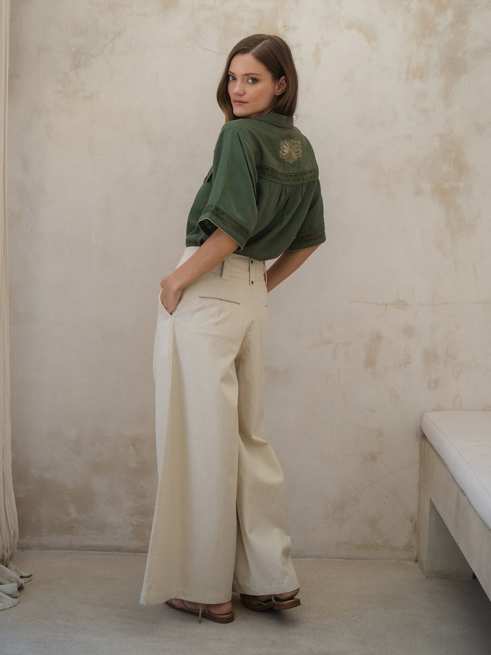Woman wearing a green shirt and white pants against a beige wall.