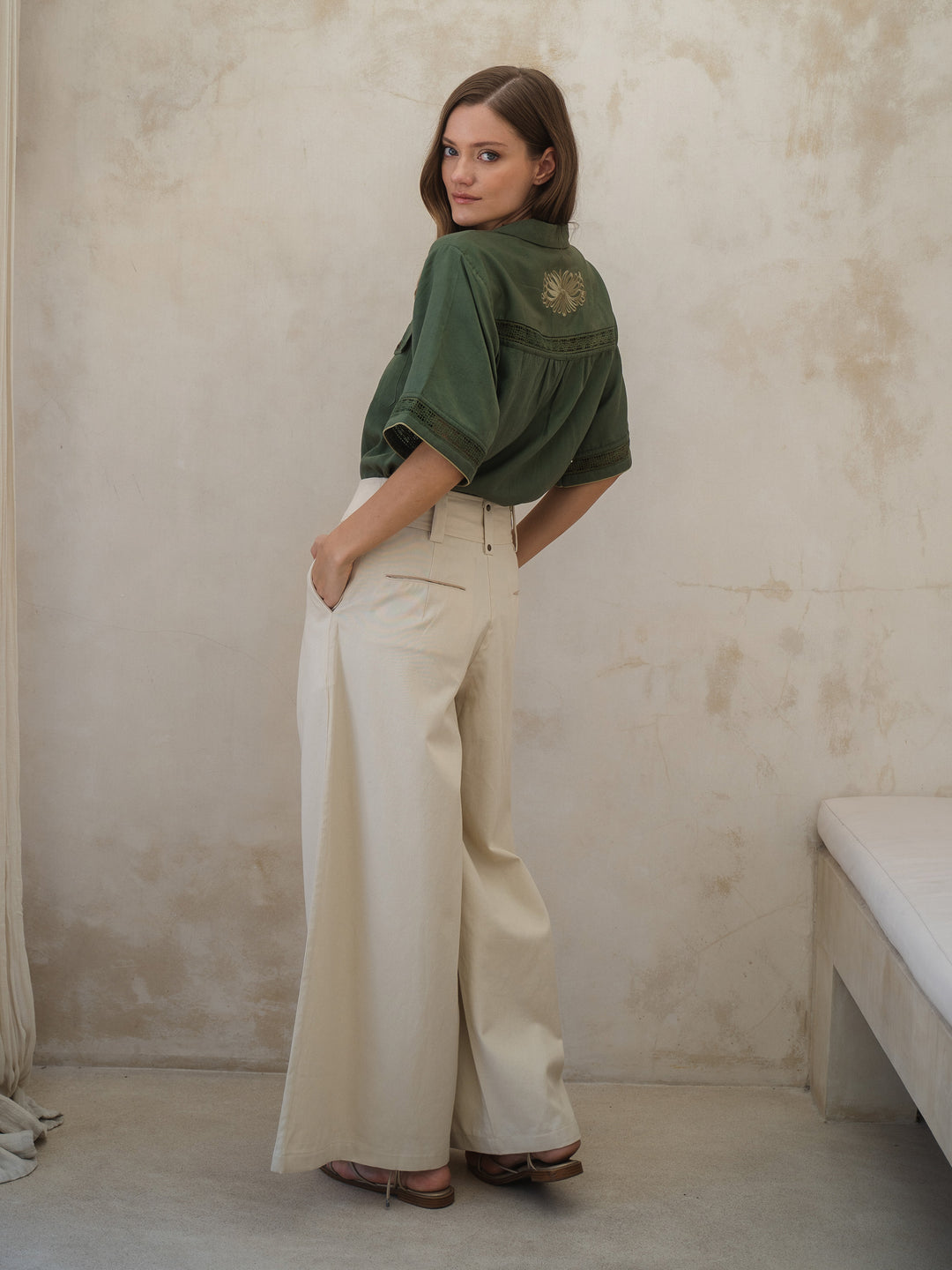 Woman wearing a green shirt and white pants against a beige wall.