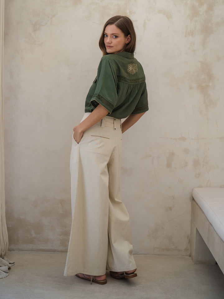 Woman wearing a green shirt and white pants against a beige wall.