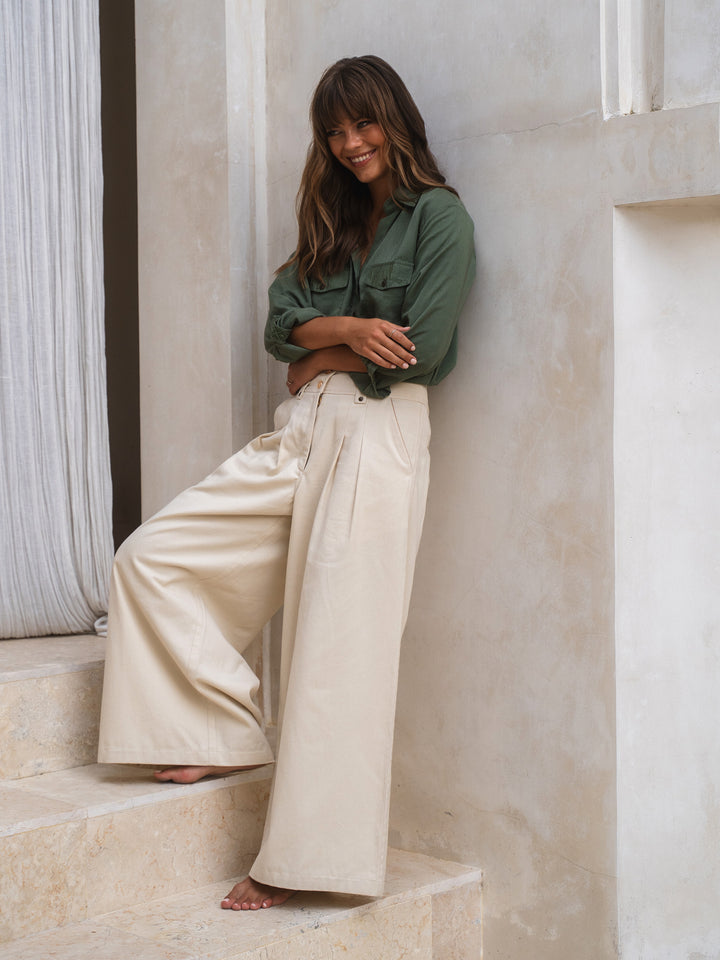 Woman in green shirt and beige pants sitting on steps with a neutral background