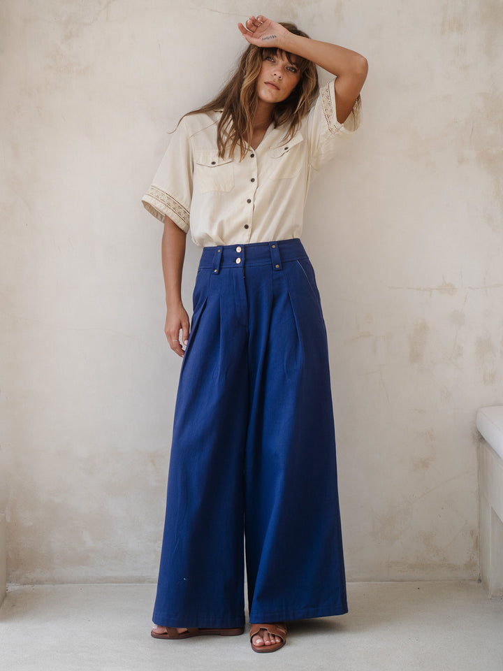 Woman wearing a beige blouse and blue wide-leg pants against a plain wall.