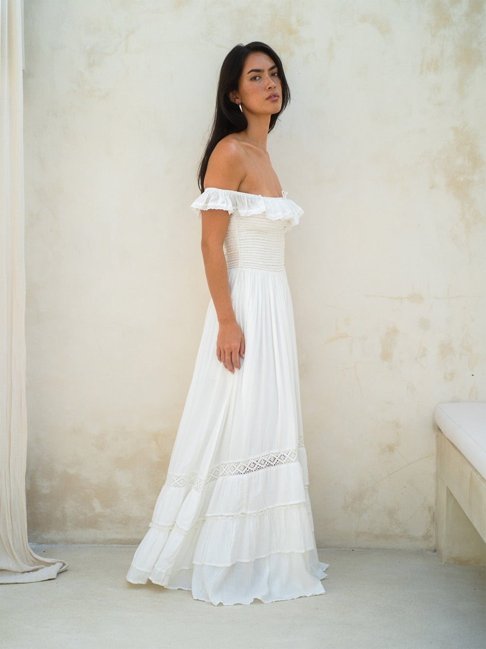 Woman wearing a white off-shoulder dress against a plain wall.