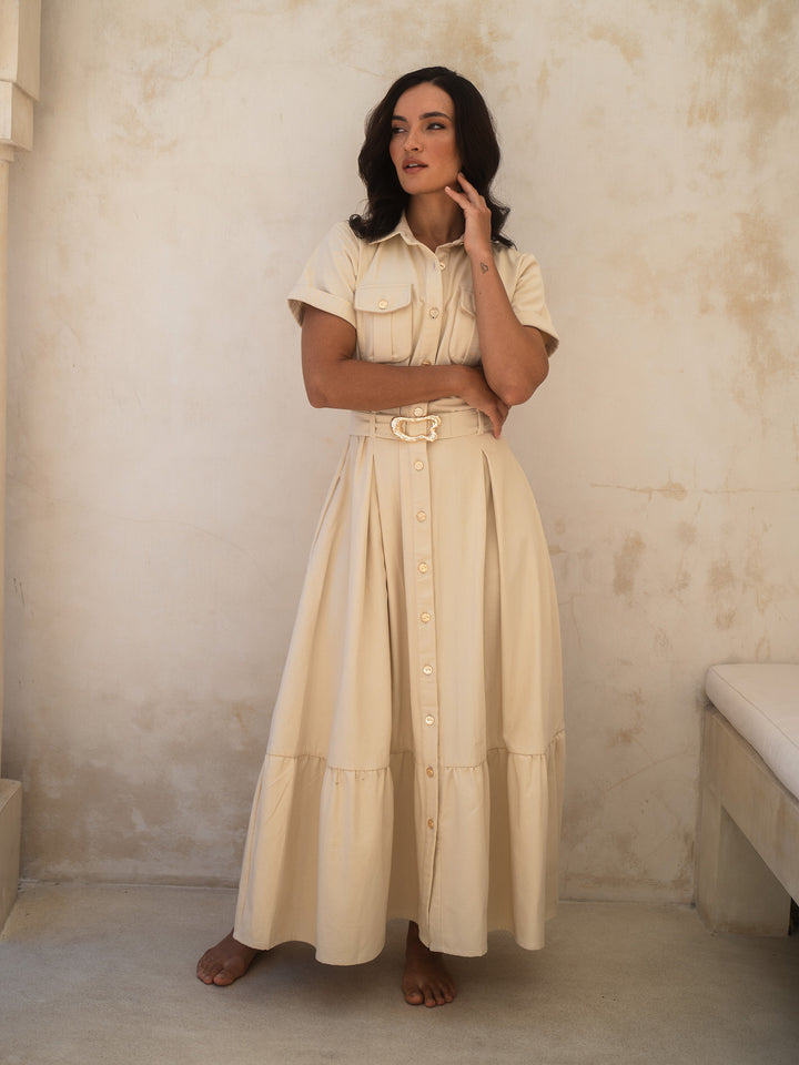 Woman wearing a beige dress with a belt against a plain wall.