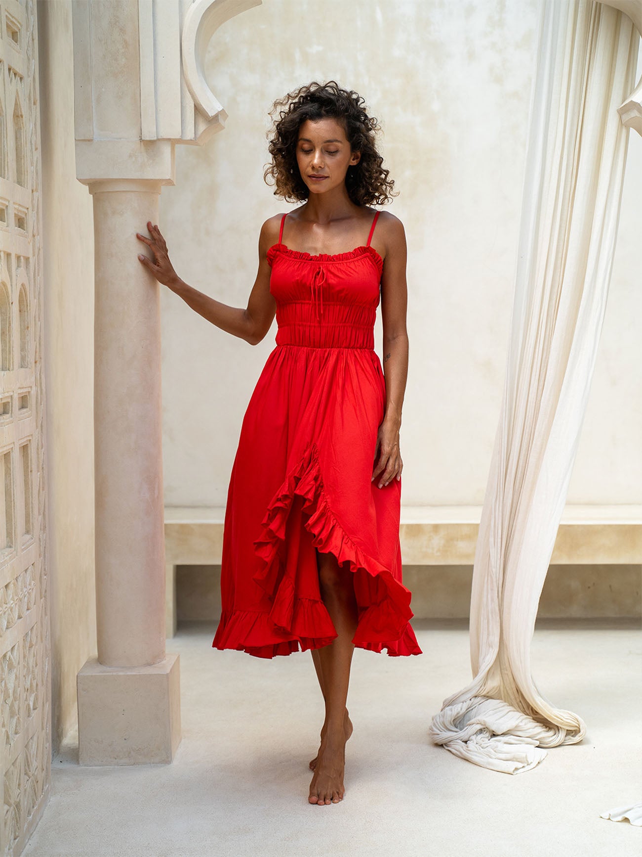 Woman wearing a red dress standing in a classical architectural setting with columns and curtains.