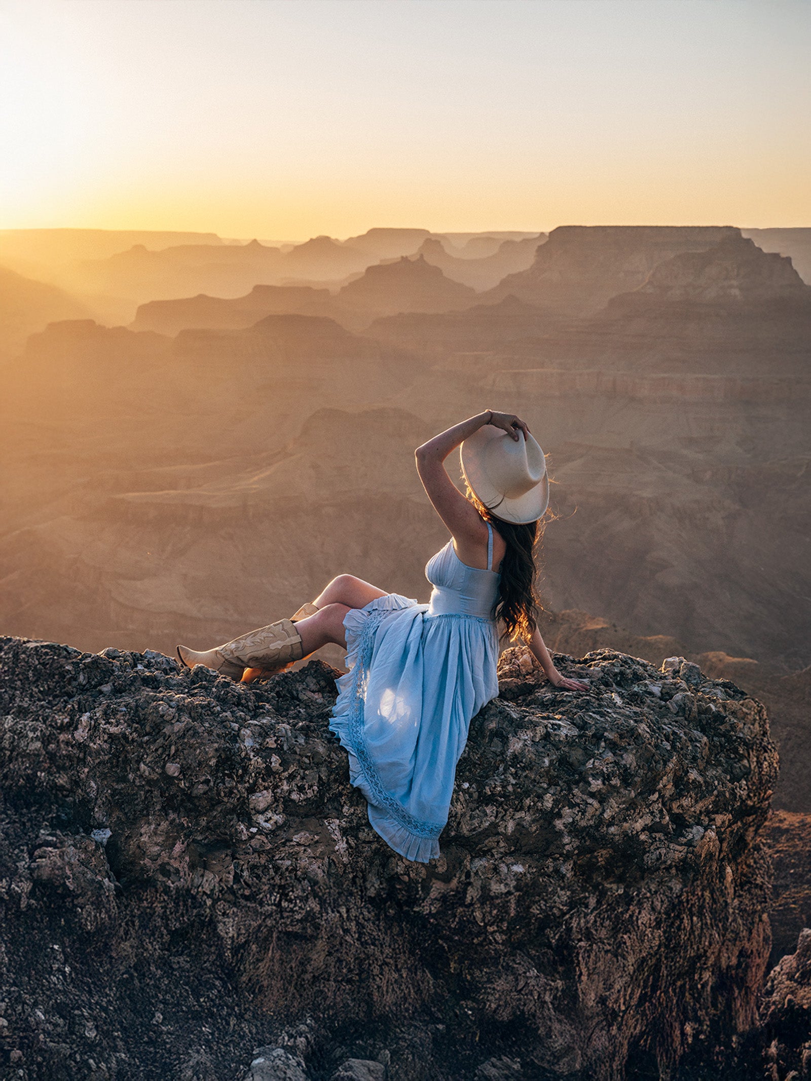 Woman in a blue dress sitting on a cliff overlooking a vast canyon at sunset.