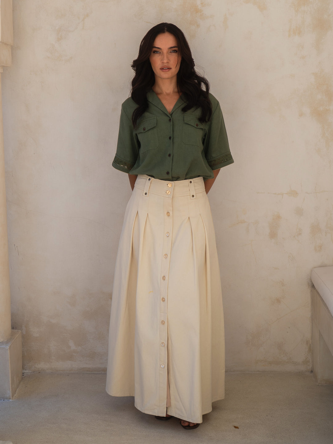 Woman wearing a green shirt and beige skirt against a textured wall.