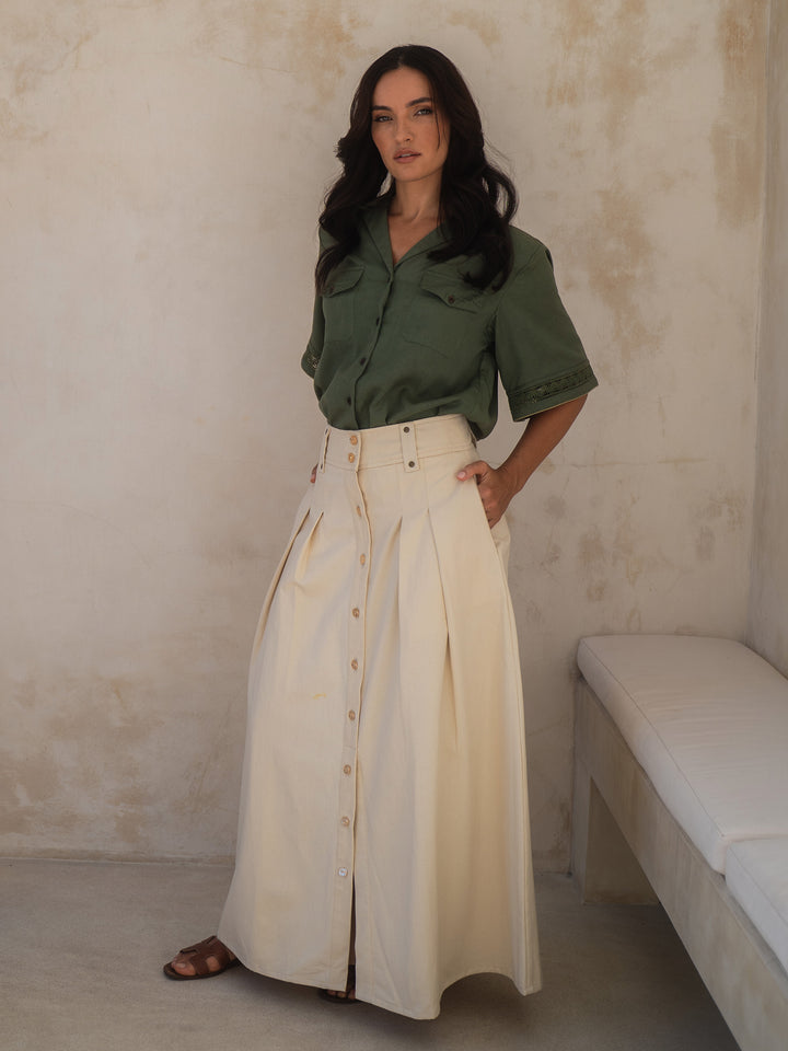Woman wearing a green shirt and beige skirt standing against a plain wall.