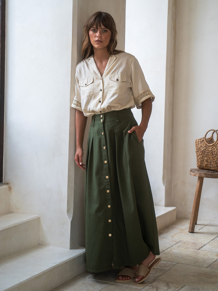 Woman wearing a beige shirt and green skirt standing in a bright room with a wooden stool and bag.