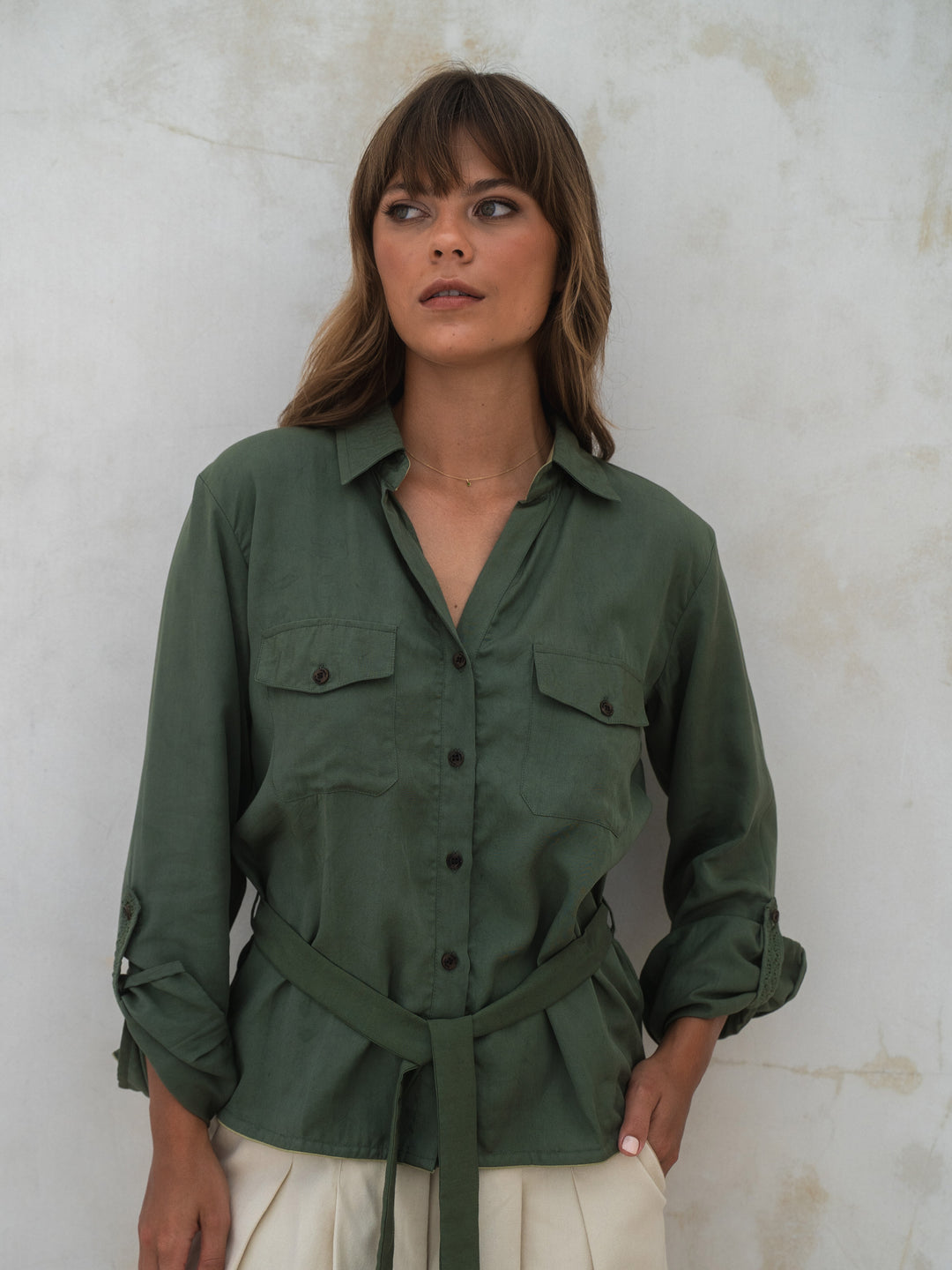 Woman wearing a green shirt with rolled-up sleeves against a textured white wall.
