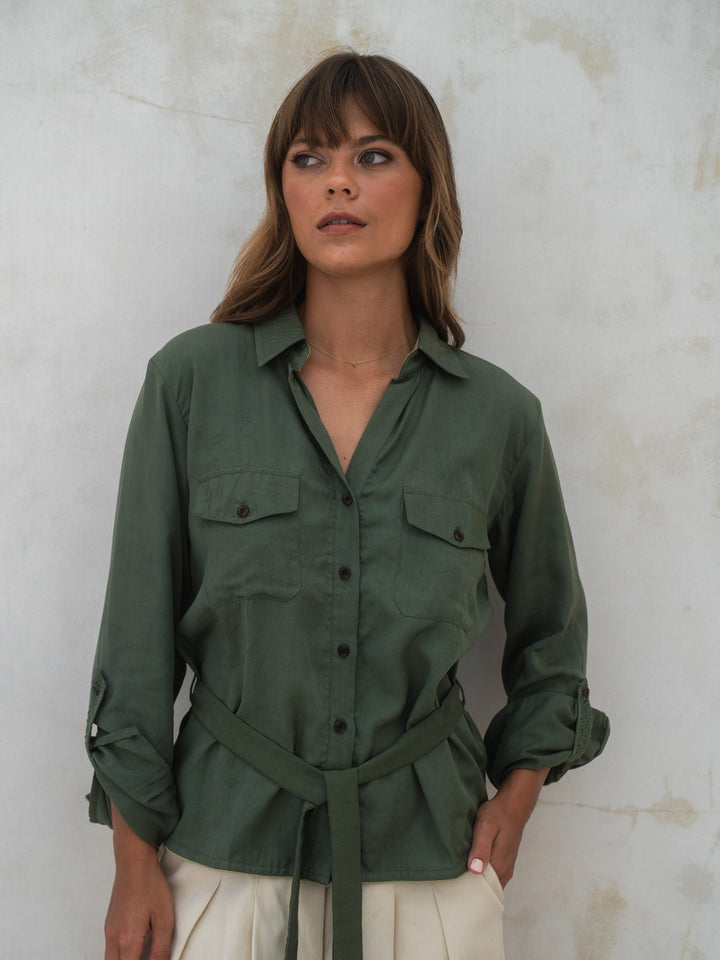 Woman wearing a green shirt with rolled-up sleeves against a textured white wall.
