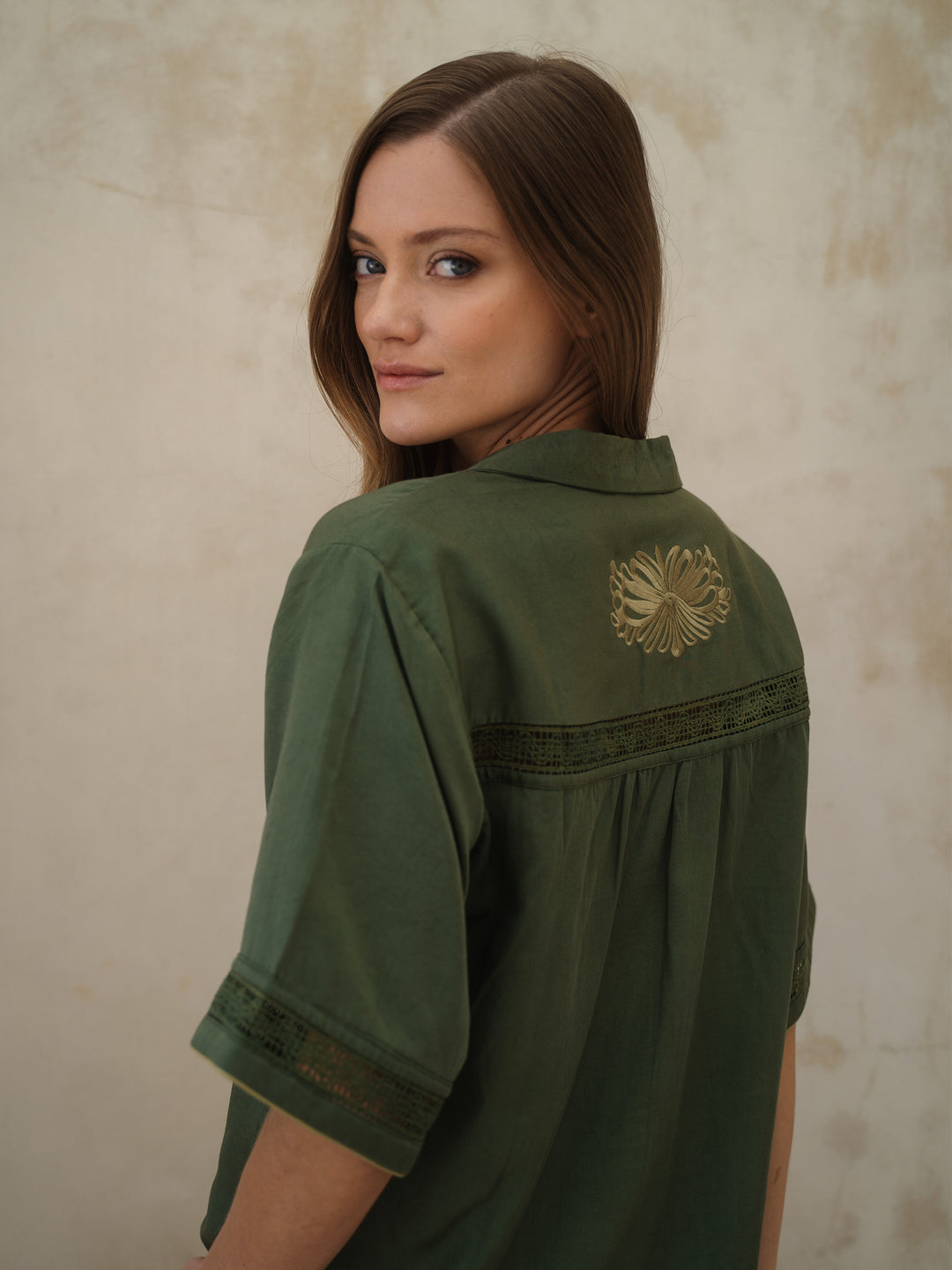 Woman wearing a green shirt with a decorative back patch against a beige wall.