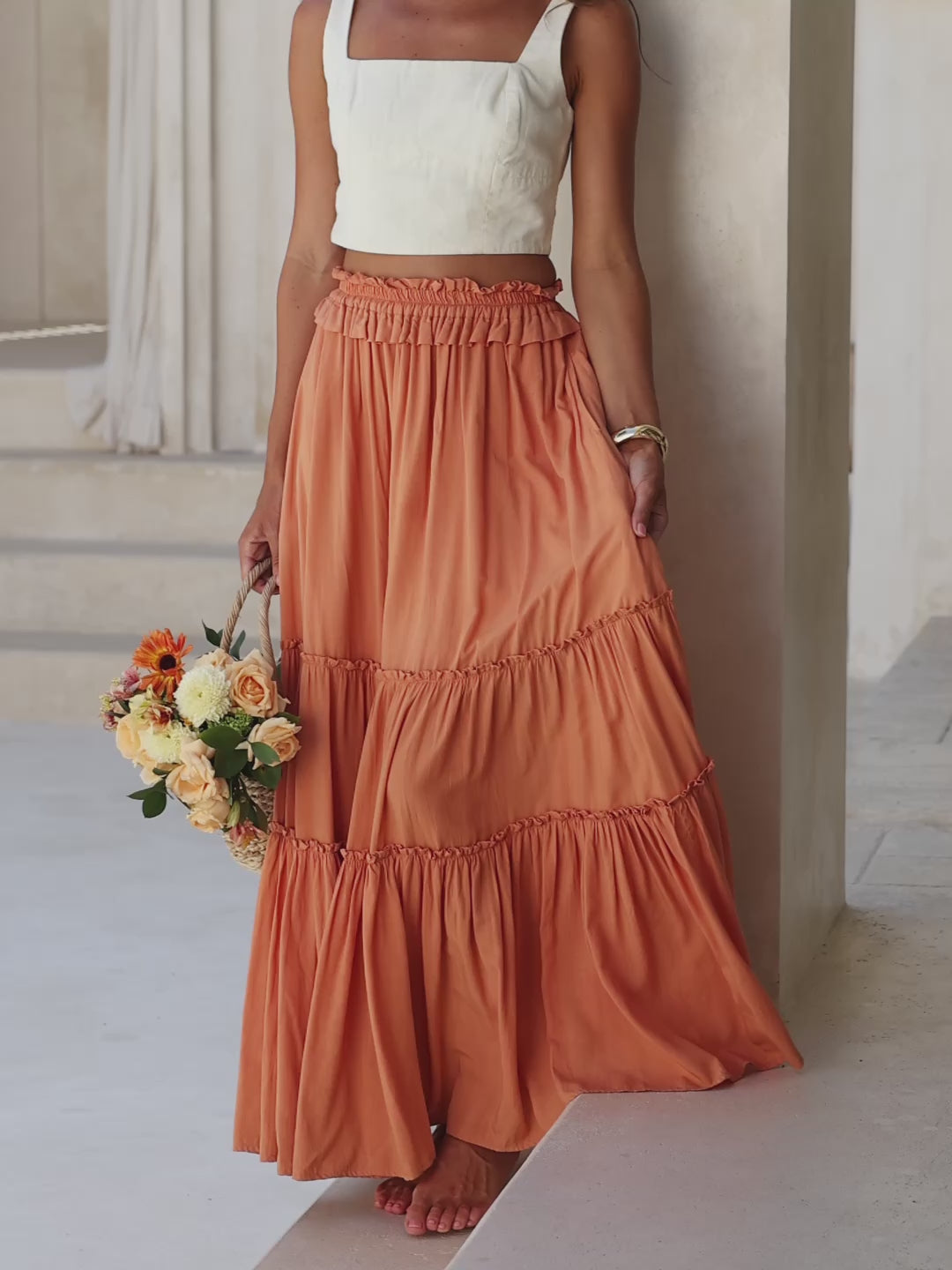 Woman in a white top and orange tiered skirt standing in a sunlit room with arched windows.