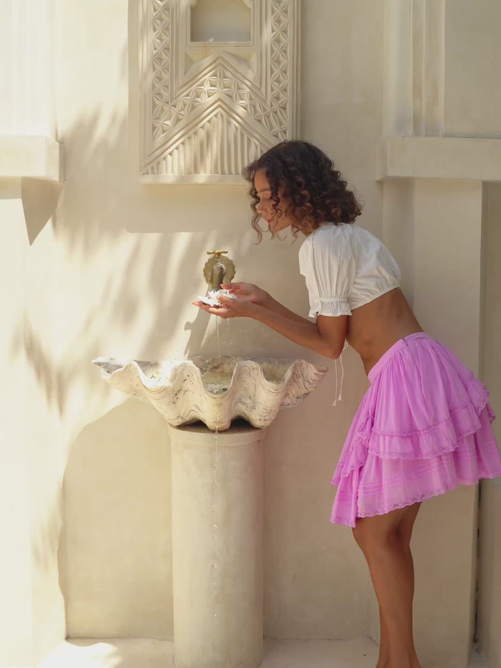 Woman wearing a white top and pink skirt standing against a neutral wall.
